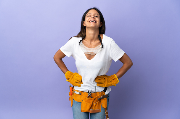 Jovem eletricista mulher isolada no fundo roxo posando com os braços no quadril e sorrindo - Foto, Imagem