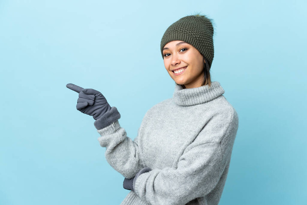Giovane ragazza uruguaiana con cappello invernale isolato su sfondo blu che punta il dito verso il lato - Foto, immagini