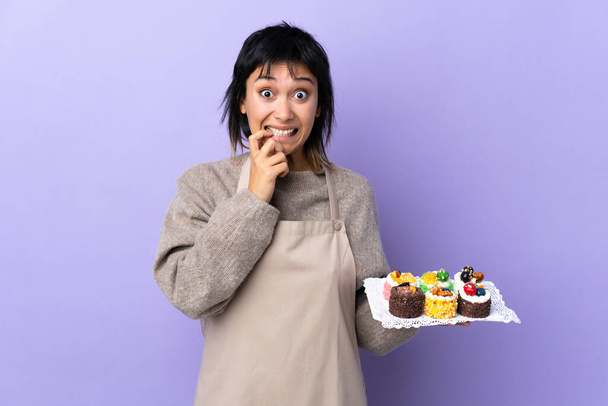 Νεαρή Ουρουγουανή γυναίκα που κρατά πολλά διαφορετικά mini cakes πάνω από απομονωμένο μωβ φόντο νευρικό και φοβισμένη - Φωτογραφία, εικόνα