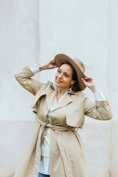 Vrouw in stijlvolle trench jas en hoed op zoek naar camera terwijl poseren buiten op straat. - Foto, afbeelding
