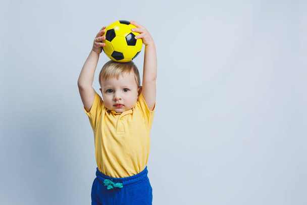Een jongetje in een geel t-shirt met een voetbal in zijn hand glimlacht geïsoleerd op een witte achtergrond. Sportkind met een bal. Sportspel voor kinderen. Kleine atleet. - Foto, afbeelding