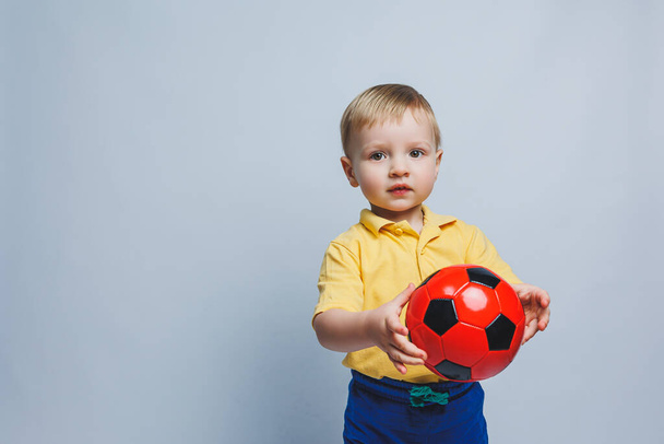 Little european boy, fan or player in yellow and blue uniform with a soccer ball, supports the soccer team on a white background. Football sport game, lifestyle concept. Isolated on white background - 写真・画像