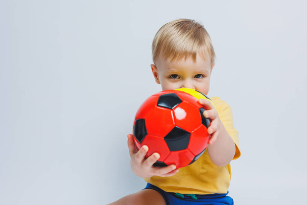 Little european boy, fan or player in yellow and blue uniform with a soccer ball, supports the soccer team on a white background. Football sport game, lifestyle concept. Isolated on white background - 写真・画像