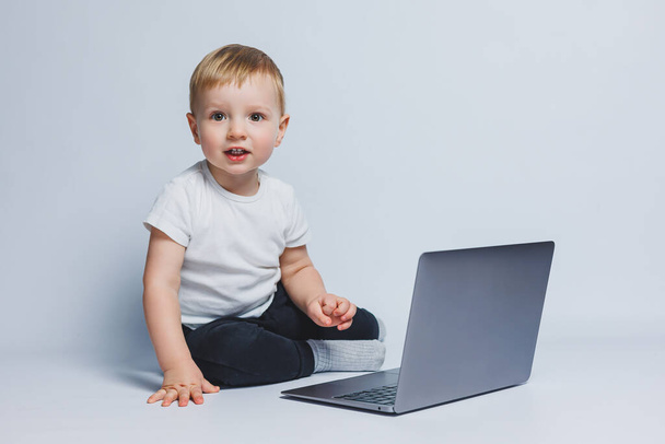 Un niño inteligente de 3-4 años se sienta con una computadora portátil sobre un fondo blanco. Un niño con una camiseta blanca y un pantalón negro se sienta en una computadora portátil y mira a la cámara. Niños modernos - Foto, Imagen