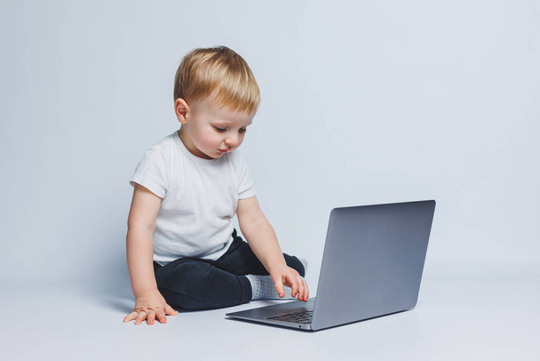 Um menino de 3-4 anos senta-se com um laptop em um fundo branco. Uma criança com uma camiseta branca e calças pretas olha para um laptop. Crianças modernas - Foto, Imagem