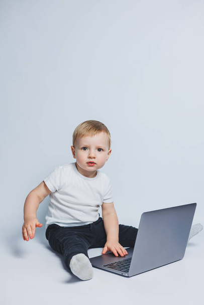 Un niño inteligente de 3-4 años se sienta con una computadora portátil sobre un fondo blanco. Un niño con una camiseta blanca y un pantalón negro se sienta en una computadora portátil y mira a la cámara. Niños modernos - Foto, Imagen