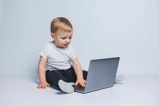 3-4 yaşlarında küçük bir çocuk beyaz arka planda dizüstü bilgisayarla oturuyor. Beyaz tişörtlü ve siyah pantolonlu bir çocuk dizüstü bilgisayara bakıyor. Modern çocuklar - Fotoğraf, Görsel