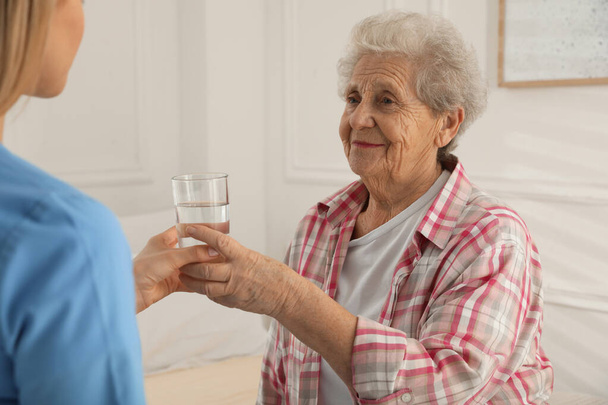 Young caregiver giving water to senior woman in room. Home care service - Fotoğraf, Görsel