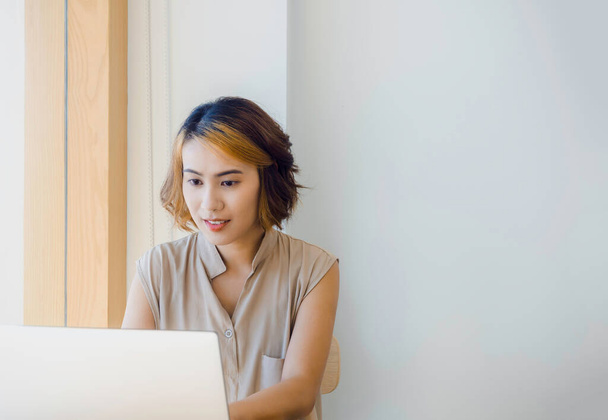 Aasialainen nainen, freelance trendikkäillä lyhyillä hiuksilla katsellen näyttöä työskennellessään kannettavan tietokoneen kanssa valkoisella seinällä taustalla kopiotila, lähellä kahvilan ikkunalasia. - Valokuva, kuva