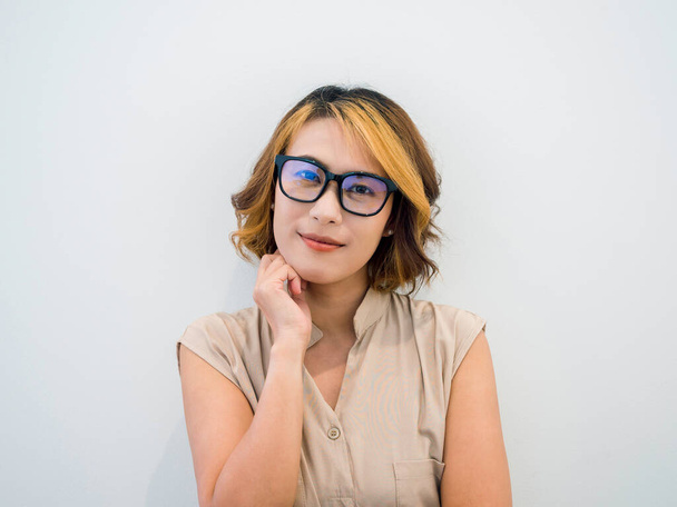 Retrato de mulher asiática sorridente cabelo curto, estilo moderno em óculos pretos e camisa bege sem mangas posando com a mão sob o queixo, olhando para a câmera no fundo branco. Trabalhando feminino com óculos. - Foto, Imagem