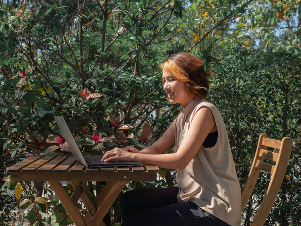 Beautiful happy Asian woman portrait with trendy short hair working, looking at a silver laptop computer screen on wooden desk with a relaxing smile in the outdoor garden. Work from anywhere. - Photo, Image