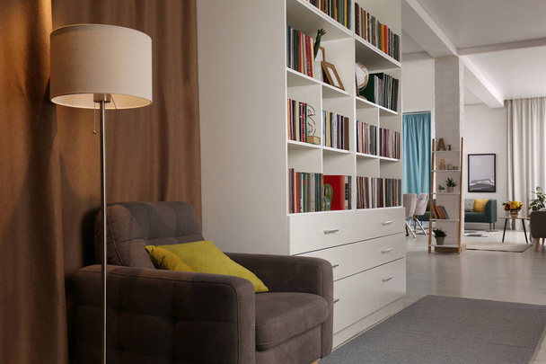 Cozy home library interior with comfortable armchair, floor lamp and collection of books on shelves - Fotó, kép