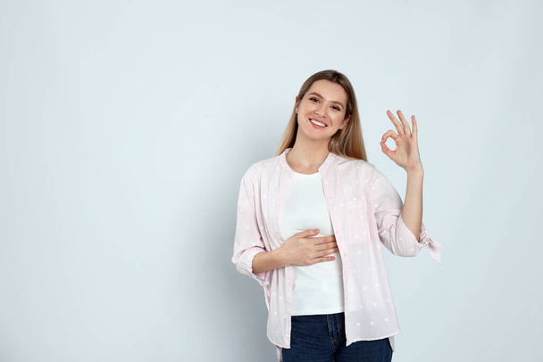 Happy woman touching her belly and showing okay gesture on light background, space for text. Concept of healthy stomach - Фото, зображення