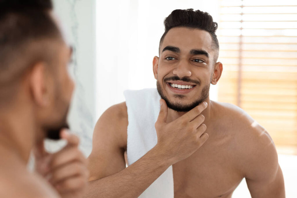 Cuidado después del afeitado. Hombre árabe guapo sin camisa con toalla en el hombro mirando en el espejo - Foto, imagen