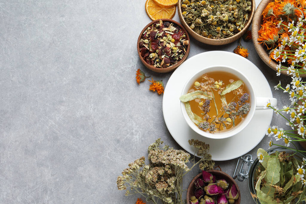 Cup of aromatic freshly brewed tea surrounded by different dry herbs on grey table, flat lay. Space for text - Photo, Image