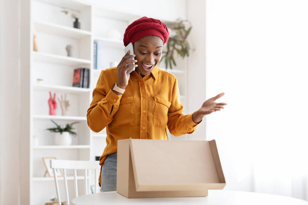 Взволнованная чёрная леди открывает коробку для доставки, разговаривает по телефону - Фото, изображение
