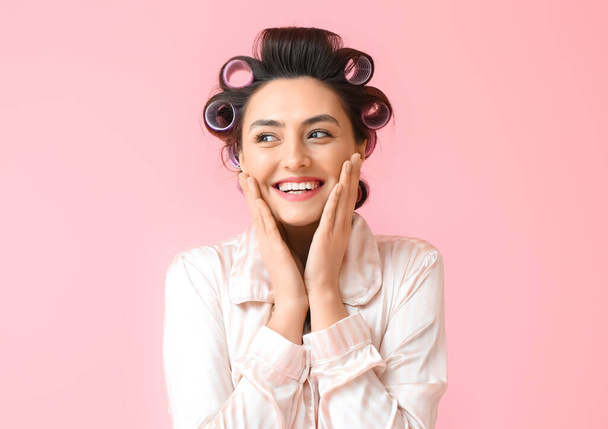 Beautiful young woman in pajamas and hair curlers on pink background - Foto, Imagem