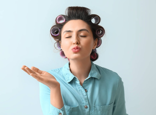 Beautiful young woman in hair rollers blowing kiss on light background - 写真・画像