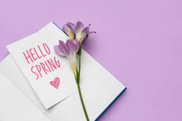 Carte avec texte HELLO SPRING, carnet et fleur de freesia sur fond lilas, gros plan - Photo, image