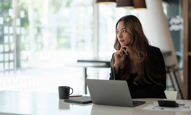 Portret van aziatische zakenvrouw zitten met laptop computer kijken buiten tijdens het werken met papierwerk in het kantoor - Foto, afbeelding