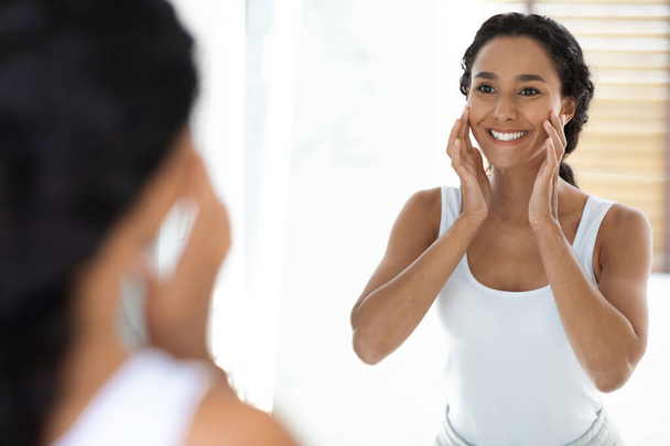 Bella sorridente arabo FemaleTouching viso mentre si guarda nello specchio in bagno - Foto, immagini