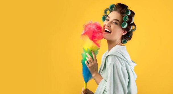 Ευτυχισμένη γυναίκα με μπούκλες μαλλιών κρατώντας φτερό ξεσκονόπανο, κίτρινο φόντο - Φωτογραφία, εικόνα