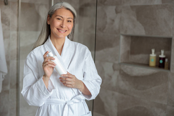 Smiling Asian lady standing in bathroom with shampoo - Φωτογραφία, εικόνα