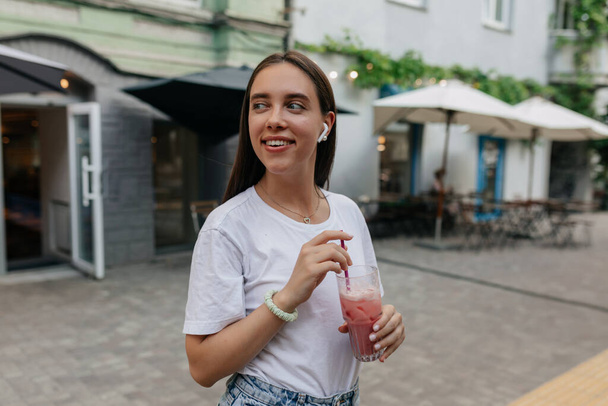 Adorable lovely girl in white t-shirt with smoothie is looking away and smiling on bright summer street in the city. Happy girl is walking on the street in good day. High quality photo - Photo, Image