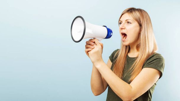Young blonde woman shouting loudly while holding a megaphone on blue background. Concept of speech and announce, idea for marketing or sales. - Foto, afbeelding