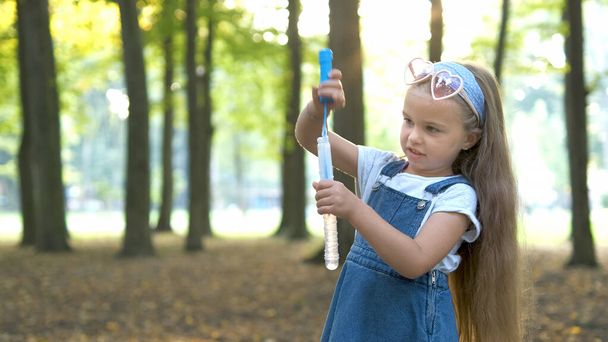 Klein gelukkig kind meisje blaast zeepbellen buiten in het groene park. Outdoor zomer activiteiten concept - Foto, afbeelding