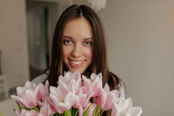 Gülümseyen ve gülümseyen mutlu kadının yakın plan portresi kameraya bakıyor ve pembe çiçekler tutuyor. Doğum gününü çiçeklerle kutlayan mutlu bayan. Yüksek kalite fotoğraf - Fotoğraf, Görsel