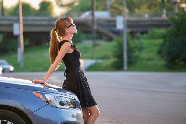 Молодая женщина-водитель наслаждается теплым летним днем, стоя рядом со своей машиной на городской улице. Концепция путешествий и отдыха - Фото, изображение