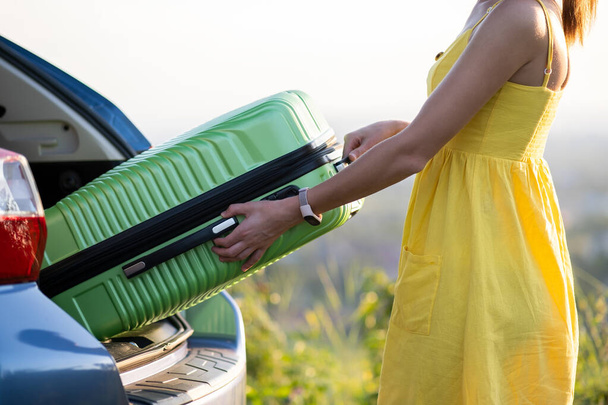 Mujer joven en vestido de verano amarillo tomando maleta verde del maletero del coche. Concepto de viajes y vacaciones - Foto, imagen
