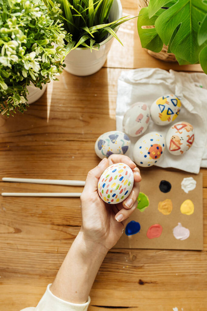 Female hand holding modern easter eggs painted. Brushes and paints with flowers and plants. Happy easter concept on a wooden background. - Photo, image