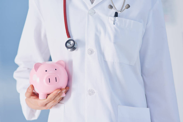 Make sure to plan for unexpected medical expenses. Cropped shot of an unrecognizable female doctor holding a piggybank while standing in the hospital. - Photo, Image
