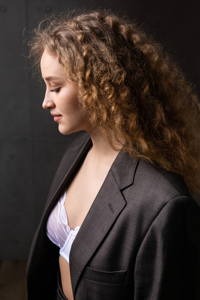 Portrait of a young red-haired curly sexy girl in a jacket with long hair in the studio. Shows off her curvaceous breasts in a bra. - Φωτογραφία, εικόνα