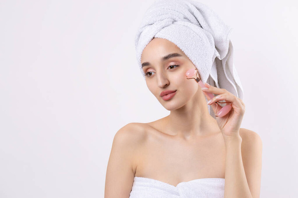 Beautiful girl with thick eyebrows and perfect skin at white background, towel on head, beauty photo. Holds a cosmetic roller. - Φωτογραφία, εικόνα