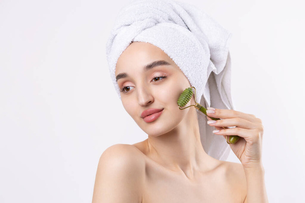 Beautiful girl with thick eyebrows and perfect skin at white background, towel on head, beauty photo. Holds a cosmetic roller. - Foto, Imagem