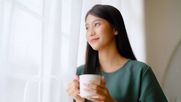 Junge asiatische Frau steht am Fenster und hält Becher im Schlafzimmer zu Hause, Sie trinkt Milch nach dem Aufwachen am Morgen - Foto, Bild