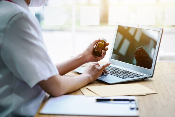 Lekarz pracujący z laptopem i trzymający butelkę lekarstwa w nowoczesnym biurze. - Zdjęcie, obraz
