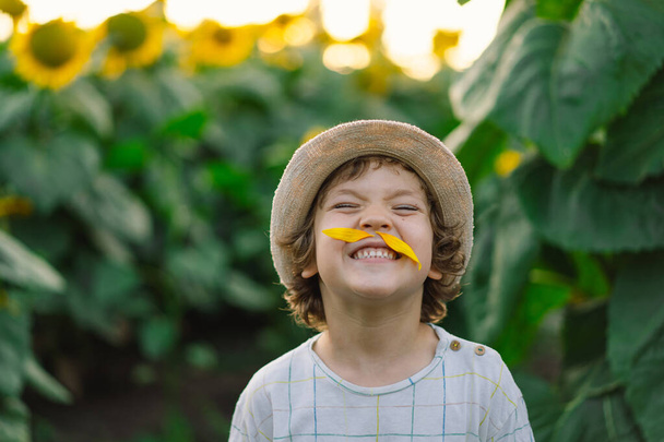 Joyeux petit garçon marchant dans le champ de tournesols et faisant une moustache à partir de pétales de tournesol. - Photo, image