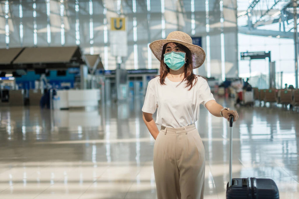 Havaalanında yürüyen yüz maskesi takan genç bir kadın, Coronavirus hastalığı enfeksiyonunu koruyan, şapkalı Asyalı bir kadın gezgin. Aşı artırıcı doz konseptinden sonra seyahat zamanı - Fotoğraf, Görsel