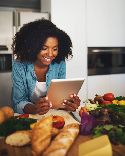 Parece que estoy haciendo todo bien. Fotografía de una joven alegre navegando en una tableta digital mientras está rodeada de verduras en la cocina en casa durante el día. - Foto, imagen