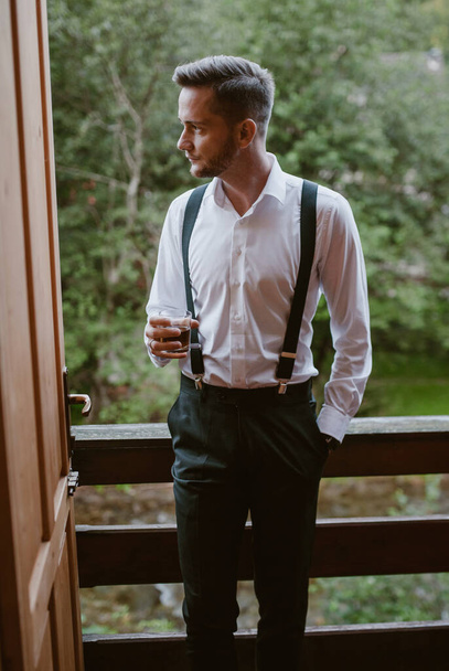 Κομψά ντυμένος γαμπρός σε ένα κοστούμι κάθεται στο μπαλκόνι ενός αγροκτήματος κούτσουρο πίνοντας ένα ποτήρι ουίσκι. Ημέρα γάμου. - Φωτογραφία, εικόνα