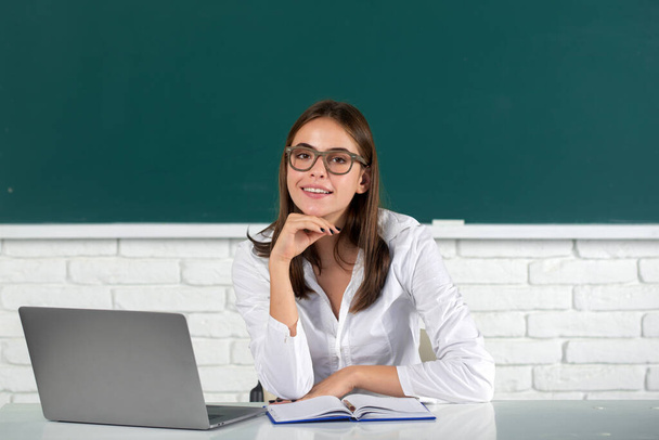 Portrait of young female college student in glasses studying in classroom on laptop in class with blackboard background. - Zdjęcie, obraz