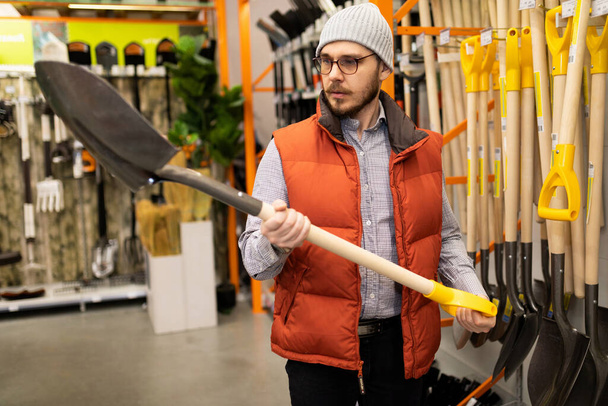 ένας αρσενικός πελάτης σε κατάστημα εργαλείων κήπου επιλέγει νέο φτυάρι για χωματουργικές εργασίες - Φωτογραφία, εικόνα