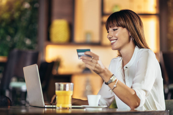 Молодая женщина сидит в кафетерии и использует кредитную карту для покупок в Интернете - Фото, изображение