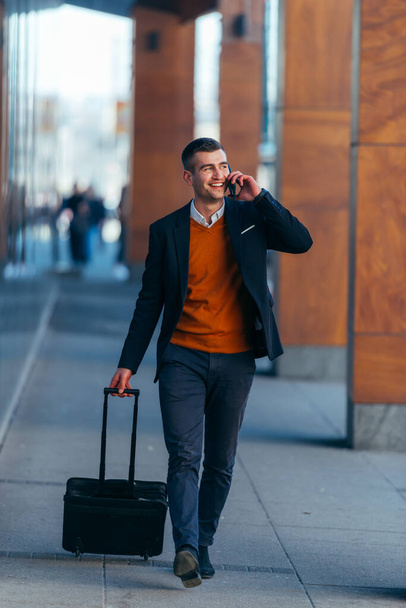 Joven hombre de negocios caminando a través de una estación mientras habla por teléfono y sacando su bolso de viaje
. - Foto, Imagen