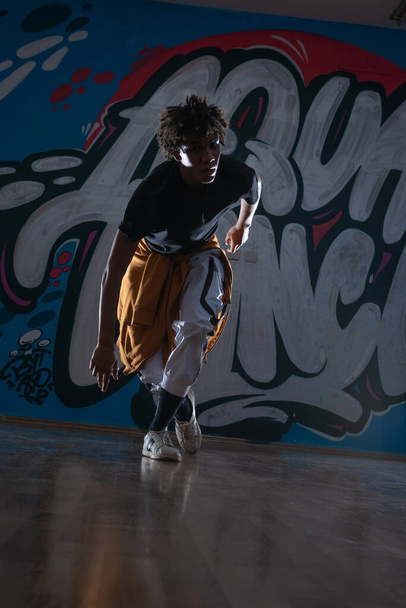 Jonge zwarte man dansend hip hop stijl in een stedelijke omgeving. hij draagt een oranje outfit en heeft een graffiti achtergrond. - Foto, afbeelding