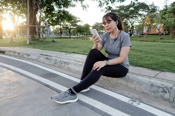 Ασιάτισσα που κάθεται στο δρόμο ακούγοντας μουσική σε ένα smartphone για να προετοιμαστεί για τρέξιμο μέσω ασύρματων ακουστικών bluetooth - Φωτογραφία, εικόνα